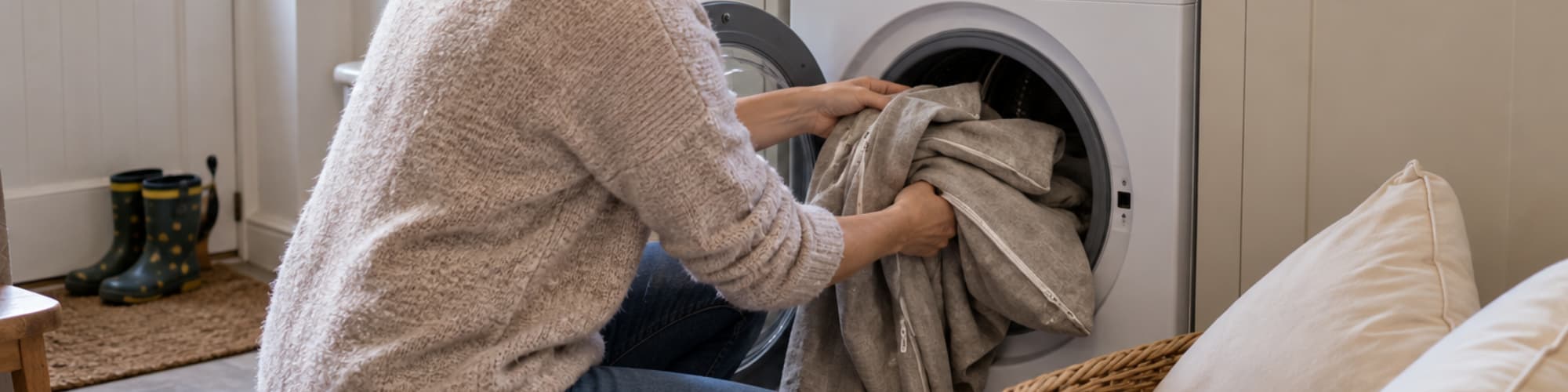 Woman loading beige sofa covers into a front-loading washing machine, with uncovered cushions in a wicker basket nearby.